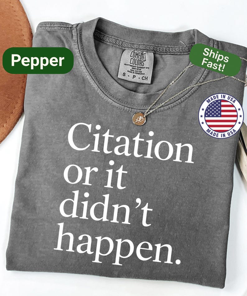 May include: A gray t-shirt with the text "Citation or it didn't happen" in white. The shirt has a crew neck and short sleeves. A silver necklace with a small pendant is draped across the shirt. The shirt is labeled "Pepper".