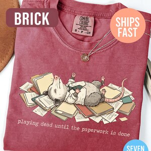 May include: Brick-colored t-shirt featuring a cartoon opossum lying on paperwork, with the text "playing dead until the paperwork is done." The shirt has a crew neck and short sleeves. A silver necklace with a pendant is visible.