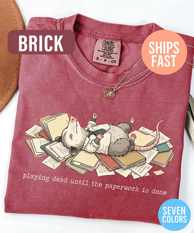May include: Brick-colored t-shirt featuring a cartoon opossum lying on paperwork, with the text "playing dead until the paperwork is done." The shirt has a crew neck and short sleeves. A silver necklace with a pendant is visible.