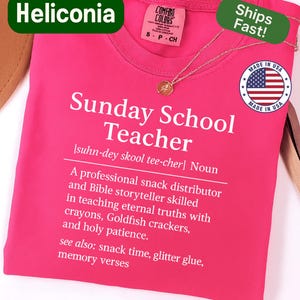 K&ouml;nnte beinhalten: Ein leuchtend pinkfarbenes T-Shirt mit dem Text "Sunday School Teacher" und einer Definition in Wei&szlig;. Eine goldene Halskette liegt auf dem T-Shirt. Der Markenname "Heliconia" wird oben links angezeigt.