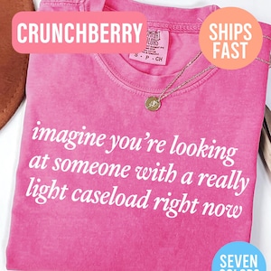 May include: A pink t-shirt with the text "imagine you're looking at someone with a really light caseload right now" in white. The shirt has a crew neck and a gold necklace with a pendant. The word "CRUNCHBERRY" is in a pink banner.