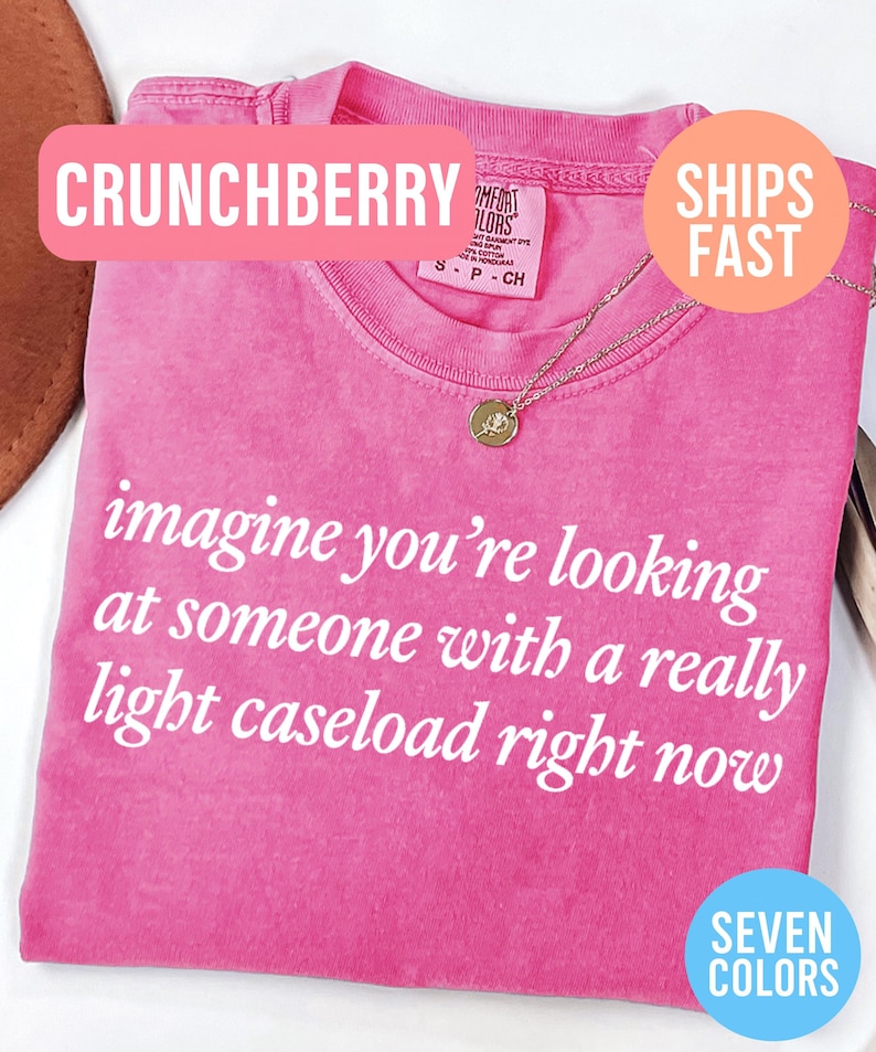 May include: A pink t-shirt with the text "imagine you're looking at someone with a really light caseload right now" in white. The shirt has a crew neck and a gold necklace with a pendant. The word "CRUNCHBERRY" is in a pink banner.