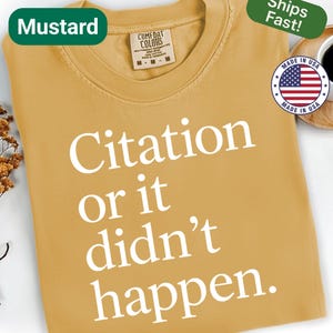 May include: Mustard-colored t-shirt with the text "Citation or it didn't happen" in white. The shirt has a crew neck and a tag that says "Comfort Colors". A green label reads "Ships Fast!" and a "Made in USA" sticker is visible.