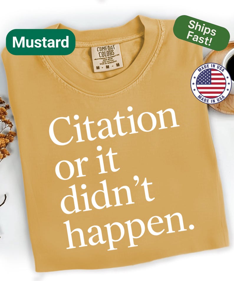 May include: Mustard-colored t-shirt with the text "Citation or it didn't happen" in white. The shirt has a crew neck and a tag that says "Comfort Colors". A green label reads "Ships Fast!" and a "Made in USA" sticker is visible.
