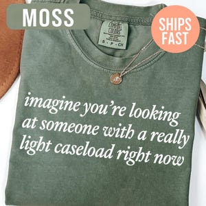 May include: Moss green t-shirt with the text "imagine you're looking at someone with a really light caseload right now" in white. A gold-colored necklace with a pendant is visible. The shirt is a Comfort Colors brand.