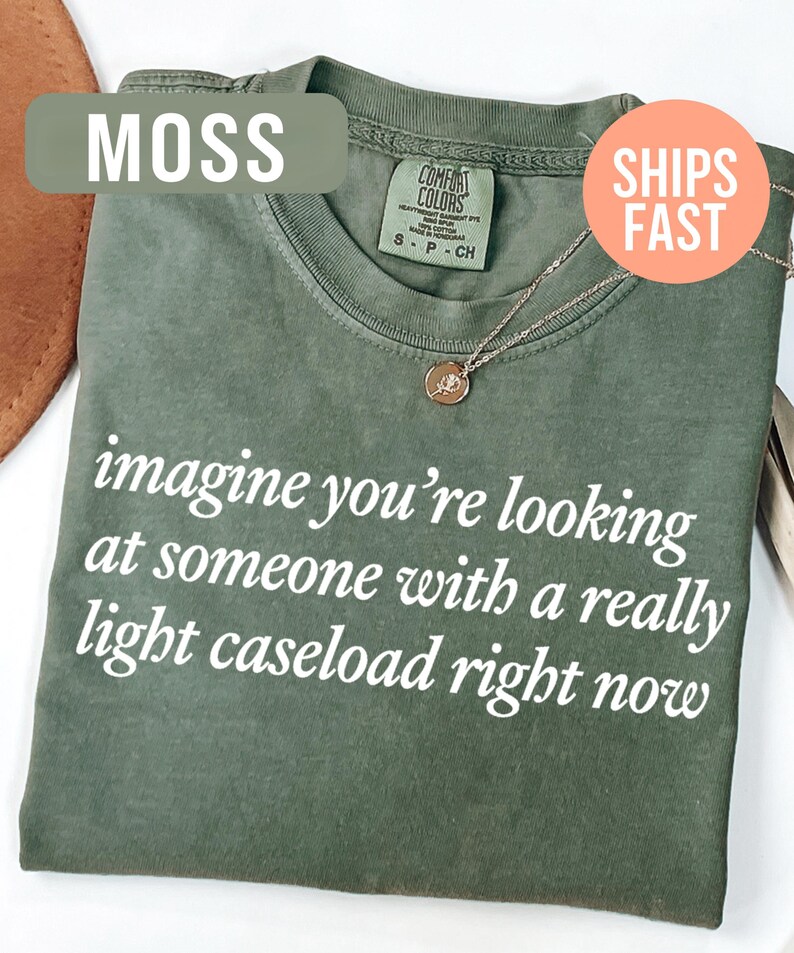 May include: Moss green t-shirt with the text "imagine you're looking at someone with a really light caseload right now" in white. A gold-colored necklace with a pendant is visible. The shirt is a Comfort Colors brand.
