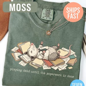 May include: Moss green t-shirt featuring a cartoon opossum lying on a pile of paperwork, wearing a suit. The text "playing dead until the paperwork is done" is printed below the illustration. A silver necklace with a pendant is visible.
