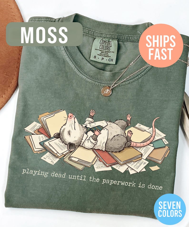 May include: Moss green t-shirt featuring a cartoon opossum lying on a pile of paperwork, wearing a suit. The text "playing dead until the paperwork is done" is printed below the illustration. A silver necklace with a pendant is visible.