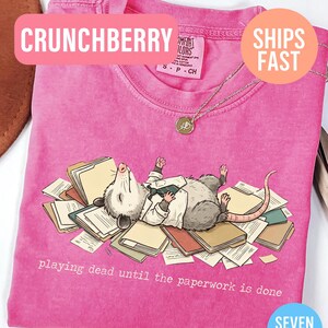 May include: A pink t-shirt with a graphic of an opossum lying on a pile of paperwork, wearing a white shirt and jacket. The text "playing dead until the paperwork is done" is below the graphic. The shirt has the text "CRUNCHBERRY" on it.