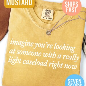 May include: Mustard-colored t-shirt with the text "imagine you're looking at someone with a really light caseload right now" in white. A silver necklace with a small pendant rests on the shirt. The shirt has a crew neck.