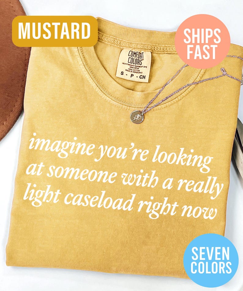 May include: Mustard-colored t-shirt with the text "imagine you're looking at someone with a really light caseload right now" in white. A silver necklace with a small pendant rests on the shirt. The shirt has a crew neck.