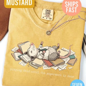 May include: Mustard-colored t-shirt with a graphic of an opossum playing dead on a pile of paperwork. The text "playing dead until the paperwork is done" is printed below the opossum. The shirt has a round neckline and short sleeves.