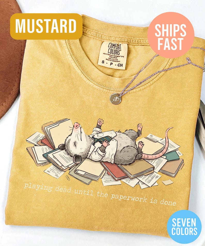 May include: Mustard-colored t-shirt with a graphic of an opossum playing dead on a pile of paperwork. The text "playing dead until the paperwork is done" is printed below the opossum. The shirt has a round neckline and short sleeves.