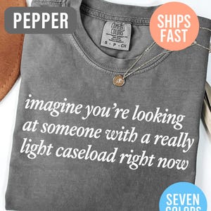 May include: A gray Comfort Colors t-shirt with the text "imagine you're looking at someone with a really light caseload right now" in white. A gold pendant necklace is visible. The shirt is labeled "Pepper" and available in seven colors.