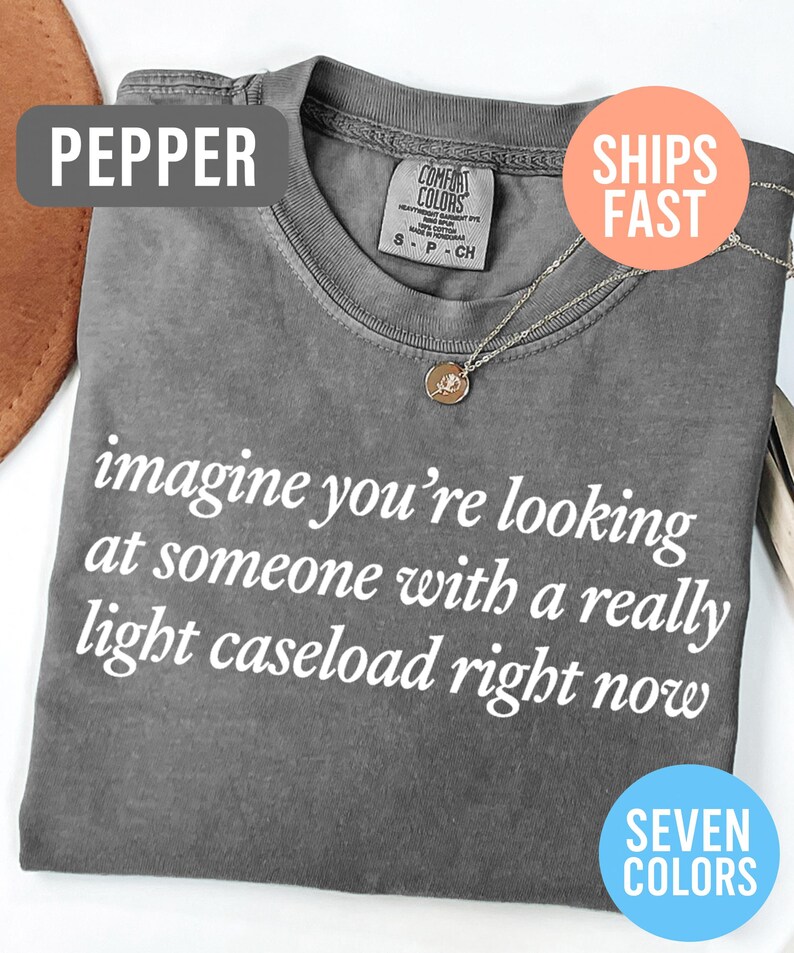 May include: A gray Comfort Colors t-shirt with the text "imagine you're looking at someone with a really light caseload right now" in white. A gold pendant necklace is visible. The shirt is labeled "Pepper" and available in seven colors.