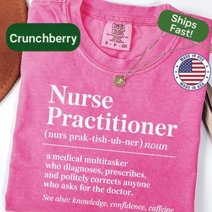 May include: A pink t-shirt with the words "Nurse Practitioner" in large white letters. The definition of a nurse practitioner is printed below. A gold necklace with a pendant rests on the shirt. The shirt has a "Made in USA" sticker.