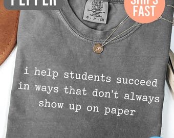 May include: Pepper-colored t-shirt with the text "i help students succeed in ways that don't always show up on paper" in white. The shirt has a crew neck and short sleeves. A gold pendant necklace is visible.