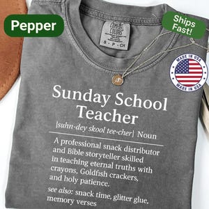 K&ouml;nnte beinhalten: Ein graues Comfort Colors T-Shirt mit dem Text "Sunday School Teacher" und einer Definition. Das Shirt hat einen Rundhalsausschnitt und kurze &Auml;rmel. Eine goldene Anh&auml;nger Halskette und ein "Made in USA" Aufkleber sind ebenfalls sichtbar.