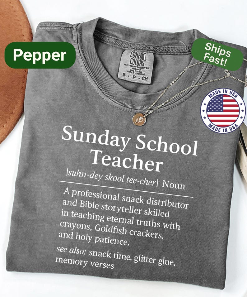 K&ouml;nnte beinhalten: Ein graues Comfort Colors T-Shirt mit dem Text "Sunday School Teacher" und einer Definition. Das Shirt hat einen Rundhalsausschnitt und kurze &Auml;rmel. Eine goldene Anh&auml;nger Halskette und ein "Made in USA" Aufkleber sind ebenfalls sichtbar.