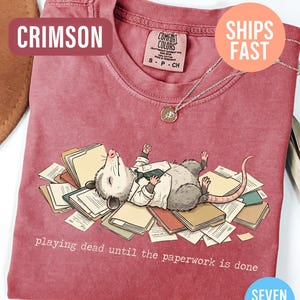 May include: Crimson-colored t-shirt featuring a cartoon opossum lying on a pile of paperwork, wearing a white shirt and tie. The text "playing dead until the paperwork is done" is printed below the opossum. The shirt has a crew neck and short sleeves.