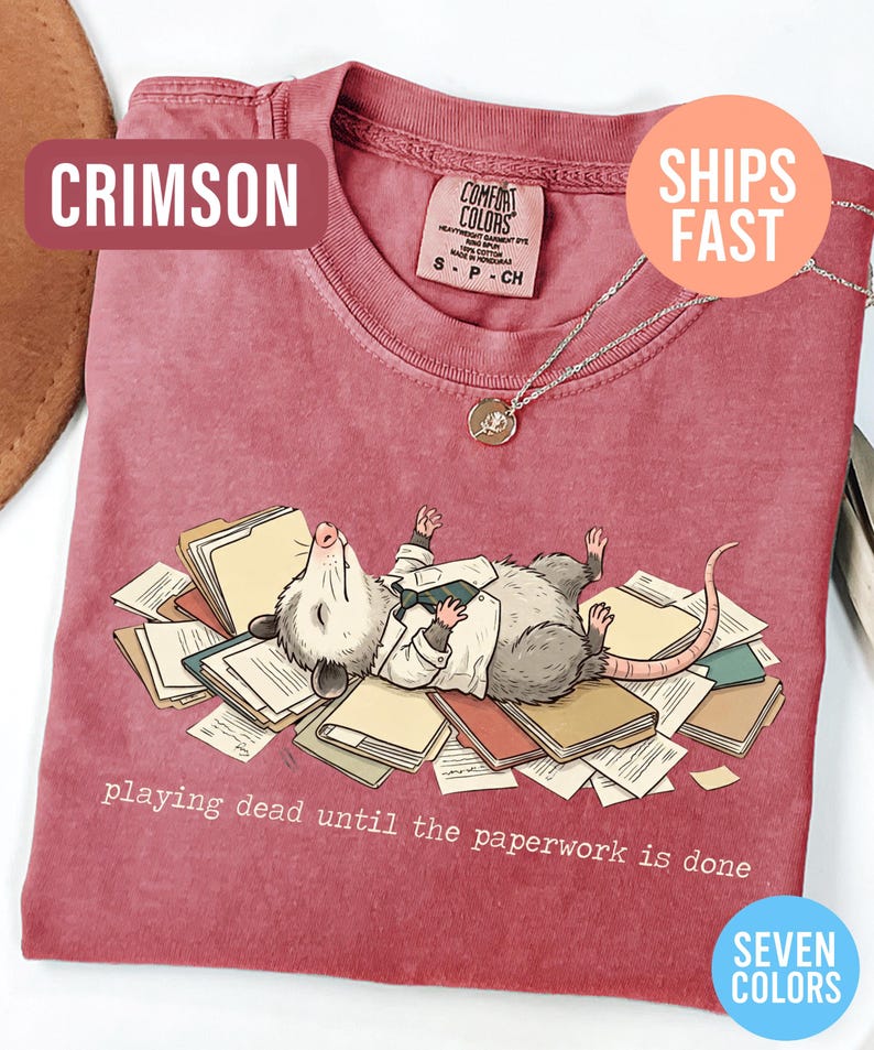 May include: Crimson-colored t-shirt featuring a cartoon opossum lying on a pile of paperwork, wearing a white shirt and tie. The text "playing dead until the paperwork is done" is printed below the opossum. The shirt has a crew neck and short sleeves.