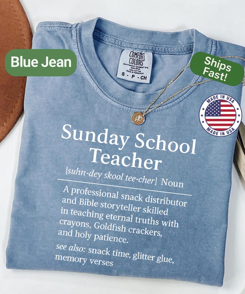 K&ouml;nnte beinhalten: Blaues Jeans-T-Shirt mit dem wei&szlig;en Aufdruck "Sunday School Teacher". Die Definition eines Sonntagsschullehrers ist ebenfalls auf dem Shirt abgedruckt. Eine goldene Halskette mit Anh&auml;nger ist &uuml;ber dem Shirt drapiert.