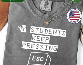 May include: A gray t-shirt with the text "MY STUDENTS KEEP PRESSING Esc" in white. The shirt has a crew neck and short sleeves. A gold-colored pendant necklace is draped on the shirt. The shirt is labeled "Pepper" and "Made in USA".