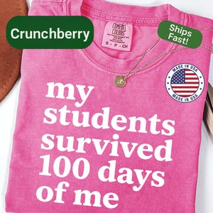 May include: A pink t-shirt with the text "my students survived 100 days of me" in white. The shirt has a crew neck and a small gold necklace. The shirt is labeled "Crunchberry" and "Made in USA".