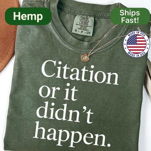 May include: Olive green t-shirt with the text "Citation or it didn't happen" in white. The shirt has a crew neck and short sleeves. A gold-colored necklace with a pendant is draped on the shirt. The shirt is labeled "Hemp" and "Made in USA".