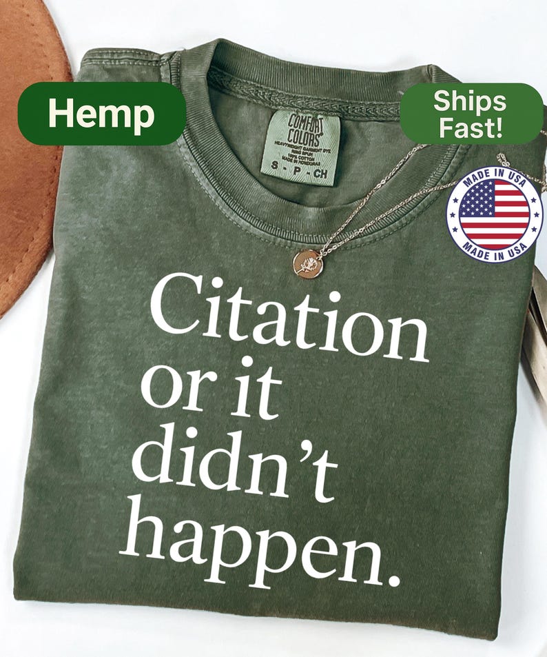 May include: Olive green t-shirt with the text "Citation or it didn't happen" in white. The shirt has a crew neck and short sleeves. A gold-colored necklace with a pendant is draped on the shirt. The shirt is labeled "Hemp" and "Made in USA".