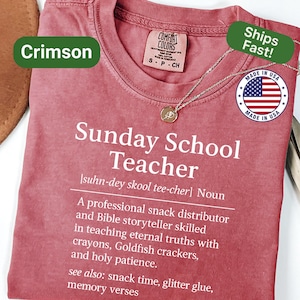 K&ouml;nnte beinhalten: Karmesinrotes T-Shirt mit dem Text "Sunday School Teacher" und einer Definition. Das Shirt hat einen Rundhalsausschnitt und kurze &Auml;rmel. Eine goldene Anh&auml;nger Halskette ist auf dem Shirt drapiert. Hergestellt in den USA.