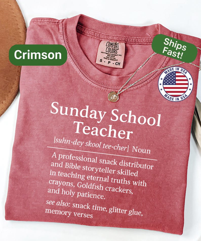 K&ouml;nnte beinhalten: Karmesinrotes T-Shirt mit dem Text "Sunday School Teacher" und einer Definition. Das Shirt hat einen Rundhalsausschnitt und kurze &Auml;rmel. Eine goldene Anh&auml;nger Halskette ist auf dem Shirt drapiert. Hergestellt in den USA.