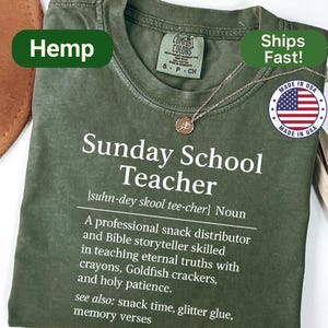 K&ouml;nnte beinhalten: Olivgr&uuml;nes T-Shirt mit dem Text "Sunday School Teacher" und einer Definition. Das Shirt hat einen Rundhalsausschnitt und kurze &Auml;rmel. Eine goldfarbene Halskette mit Anh&auml;nger ist sichtbar. Das Shirt ist mit "Hemp" und "Made in USA" gekennzeichnet.