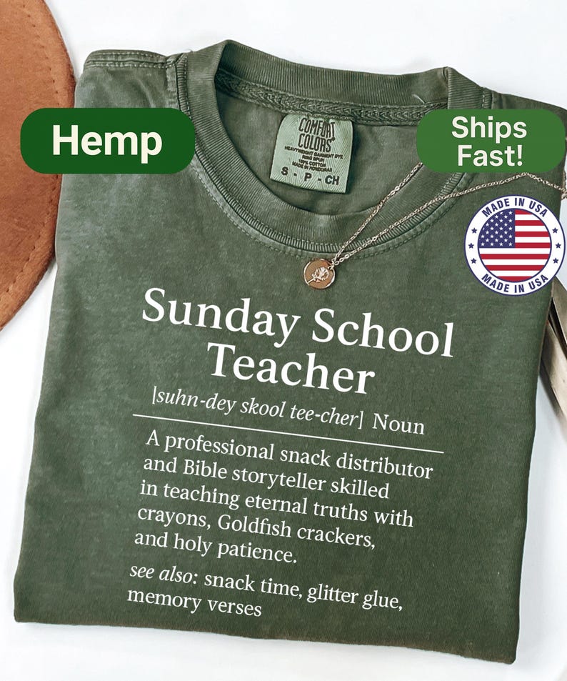 K&ouml;nnte beinhalten: Olivgr&uuml;nes T-Shirt mit dem Text "Sunday School Teacher" und einer Definition. Das Shirt hat einen Rundhalsausschnitt und kurze &Auml;rmel. Eine goldfarbene Halskette mit Anh&auml;nger ist sichtbar. Das Shirt ist mit "Hemp" und "Made in USA" gekennzeichnet.