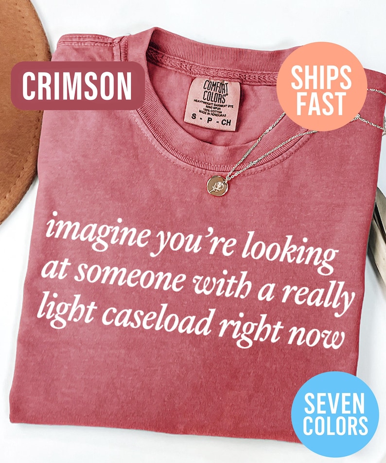 May include: Crimson-colored t-shirt with the text "imagine you're looking at someone with a really light caseload right now." The shirt has a crew neck and a small pendant necklace. The image also includes the text "Seven Colors."