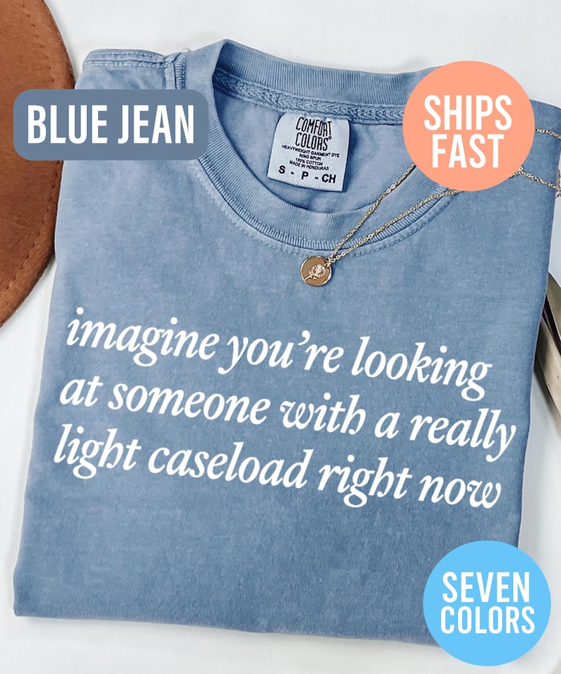 May include: Blue jean colored t-shirt with the text "imagine you're looking at someone with a really light caseload right now" in white. A gold necklace with a pendant is on the shirt. The shirt is labeled "Comfort Colors".