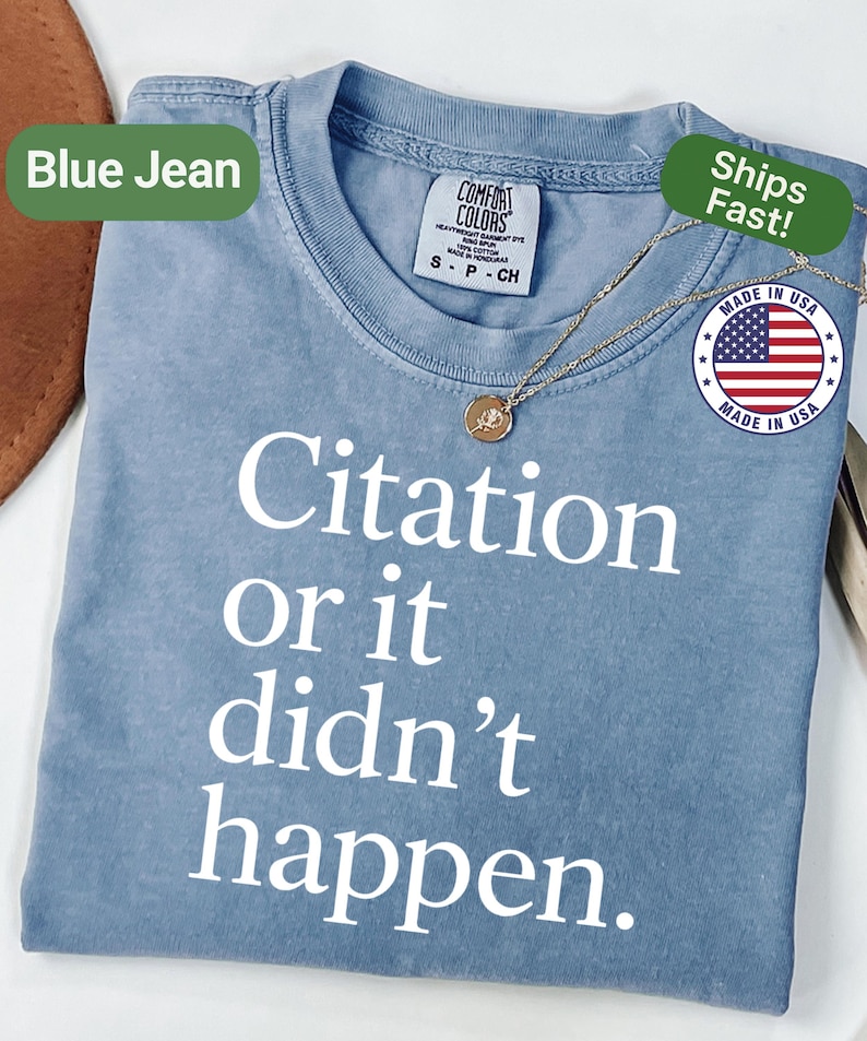May include: A blue jean colored t-shirt with the text "Citation or it didn't happen" in white. The shirt has a crew neck and short sleeves. A gold necklace with a pendant is draped on the shirt. The shirt has a "Made in USA" tag.