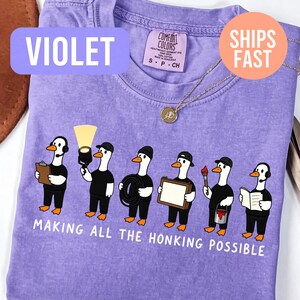 May include: A violet t-shirt featuring a graphic of seven cartoon geese dressed in black crew uniforms, each holding stage equipment like a clipboard, spotlight, cable, frame, paintbrush, and headset, with the text "MAKING ALL THE HONKING POSSIBLE."