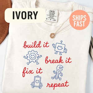 May include: An ivory t-shirt featuring red text that reads build it, break it, fix it, repeat. The design includes blue cartoon illustrations of a square robot, a gear, a battery, and a wrench, each with a smiling face and limbs.