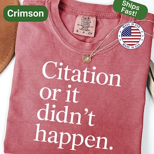 May include: Crimson-colored t-shirt with the text "Citation or it didn't happen" in white. The shirt has a crew neck and short sleeves. A gold-colored pendant necklace is draped on the shirt. The shirt is labeled "Comfort Colors" and "Made in USA".