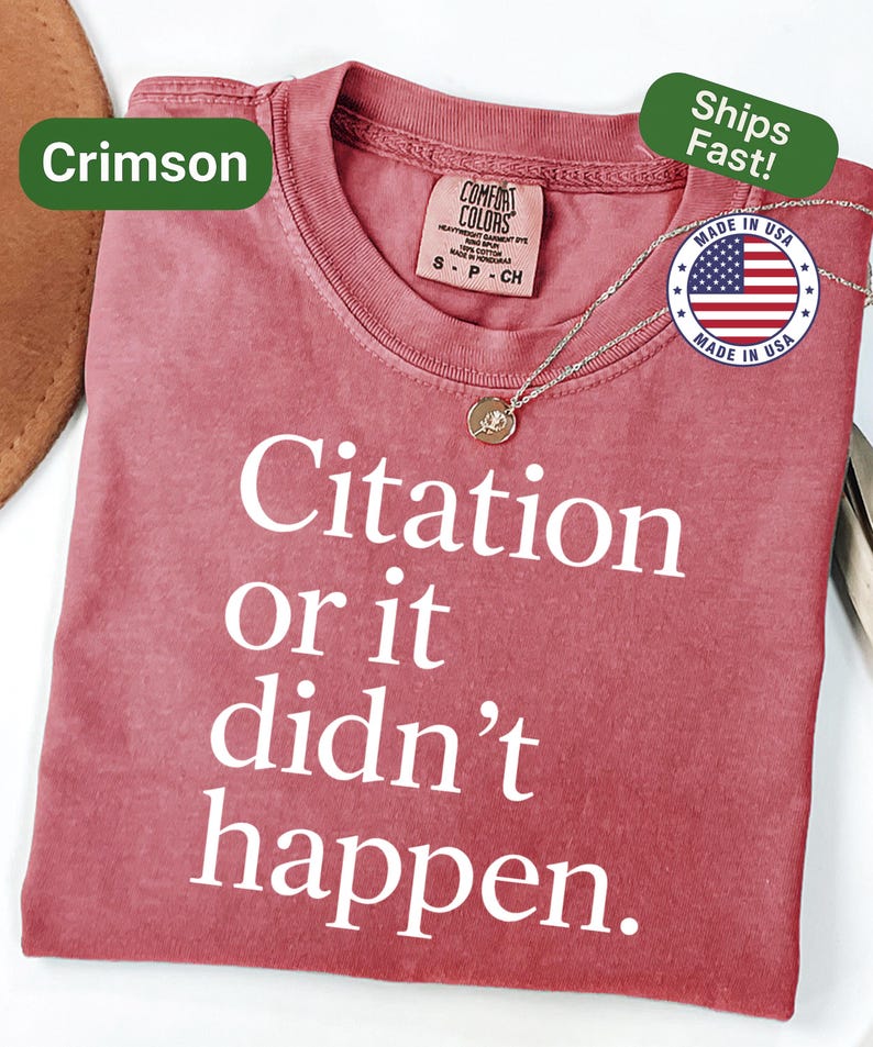 May include: Crimson-colored t-shirt with the text "Citation or it didn't happen" in white. The shirt has a crew neck and short sleeves. A gold-colored pendant necklace is draped on the shirt. The shirt is labeled "Comfort Colors" and "Made in USA".