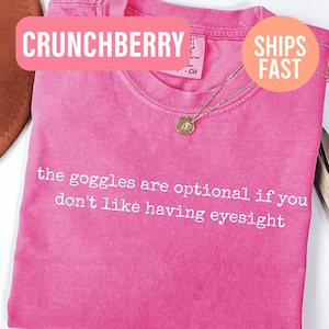 May include: A bright pink short-sleeved t-shirt with a crew neckline. The shirt features white text printed across the chest that reads: the goggles are optional if you don't like having eyesight. A gold-toned circular pendant necklace rests on top.