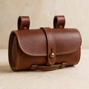 May include: A brown leather cylindrical bag featuring a front flap secured by a single brass stud closure. The bag includes two vertical leather straps on the top for attachment and a horizontal leather handle at the base with brass hardware accents.