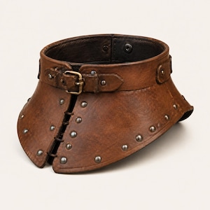 May include: A brown leather neck collar featuring a front buckle closure, dark metal studs along the edges, and a vertical lacing detail at the centre. The interior is lined with a dark material and includes a visible snap button fastener.