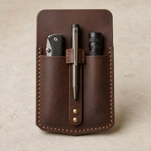 Könnte beinhalten: Ein dunkelbrauner Leder-Taschenorganizer mit zwei Hauptfächern und einer zentralen Stiftschlaufe. Der Organizer enthält ein schwarzes Klappmesser, einen metallischen Stift und eine schwarze Taschenlampe. Mit sichtbaren beigen Nähten und zwei Messingnieten.
