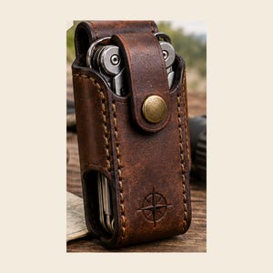 May include: A dark brown leather sheath with visible stitching and a brass snap button closure. The case contains a silver multi-tool and features an embossed compass rose design on the front flap. The leather has a textured, weathered finish.