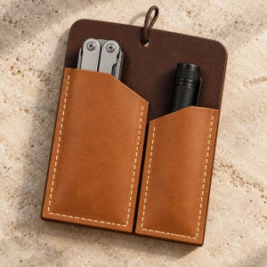 Könnte beinhalten: Eine dunkelbraune Lederhülle mit zwei hellbraunen Ledertaschen und weißen Nähten. Die linke Tasche enthält ein silbernes Multifunktionswerkzeug, die rechte eine schwarze zylindrische Taschenlampe. Oben befindet sich eine kleine Lederschlaufe.