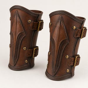 May include: A pair of dark brown leather bracers featuring a raised, pointed central ridge design. Each bracer is secured with two adjustable leather straps equipped with brass-toned metal buckles and is accented with matching brass-toned rivets.