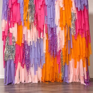 May include: A decorative backdrop composed of numerous vertical hanging strips in shades of bright pink, orange, light pink, and lavender. The display features interspersed sections of shimmering silver metallic fringe and curly purple ribbons.