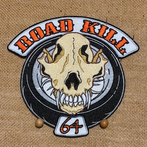 Peut inclure: Un écusson brodé représentant un crâne d'animal beige centré sur un motif de pneu noir. Le texte ROAD KILL est arqué en orange au-dessus du crâne, et le nombre 64 est affiché en orange sur une languette blanche au bas du motif circulaire.